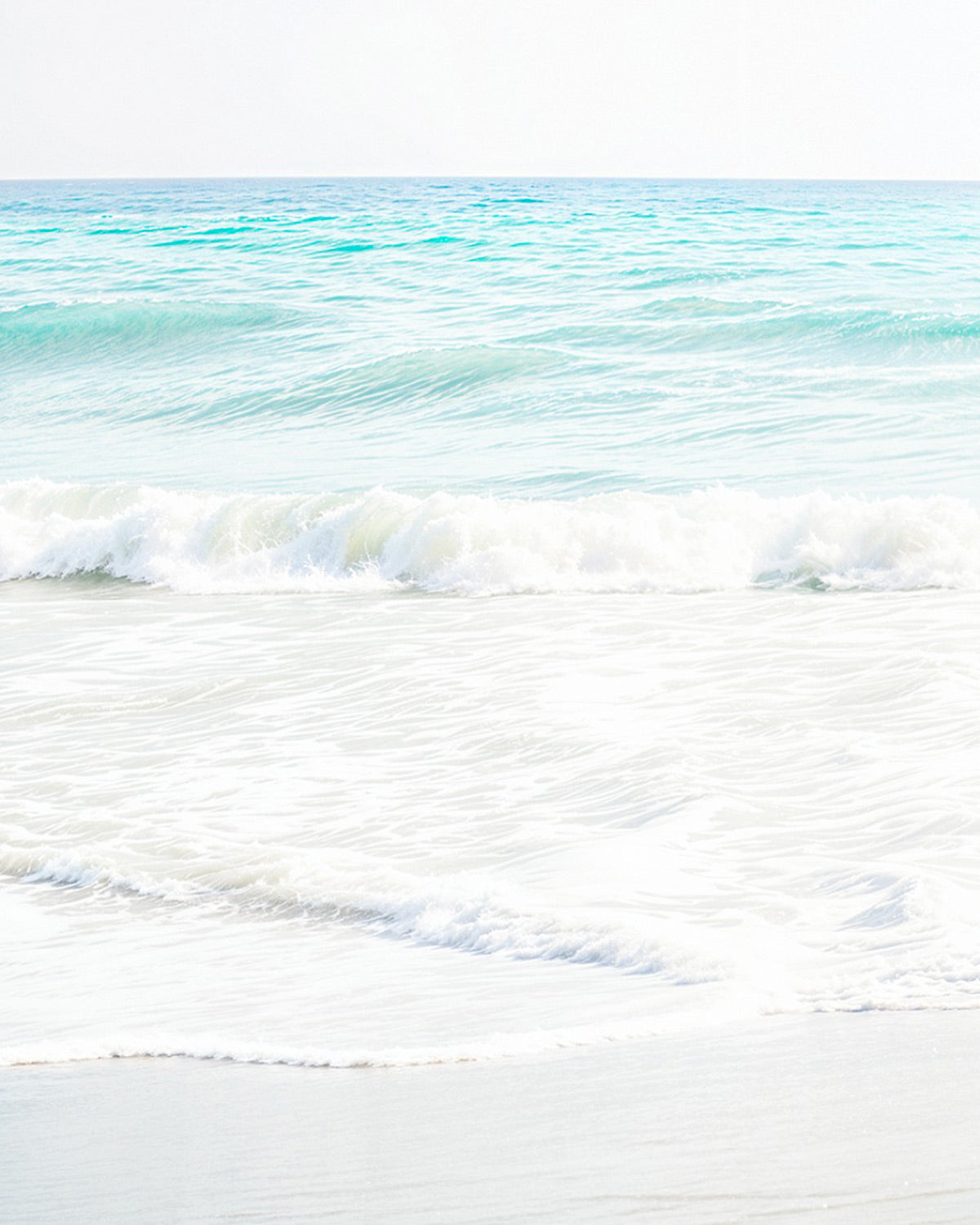 Light, airy photograph of white foamy waves on a pale sandy beach with turquoise sea behind