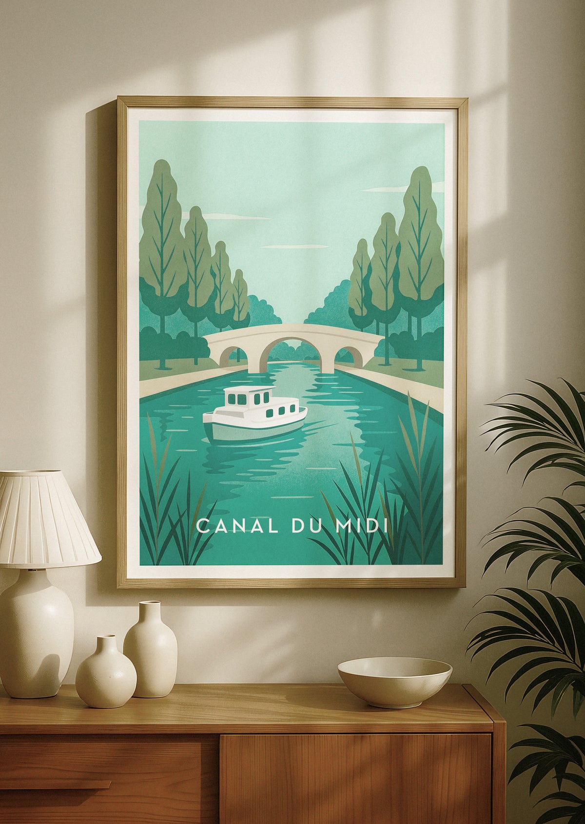 A framed travel-style poster print of the Canal du Midi in France, featuring a retro boat on teal water with trees and a stone bridge, displayed on a beige wall above a wooden console table with decorative vases and a lamp.